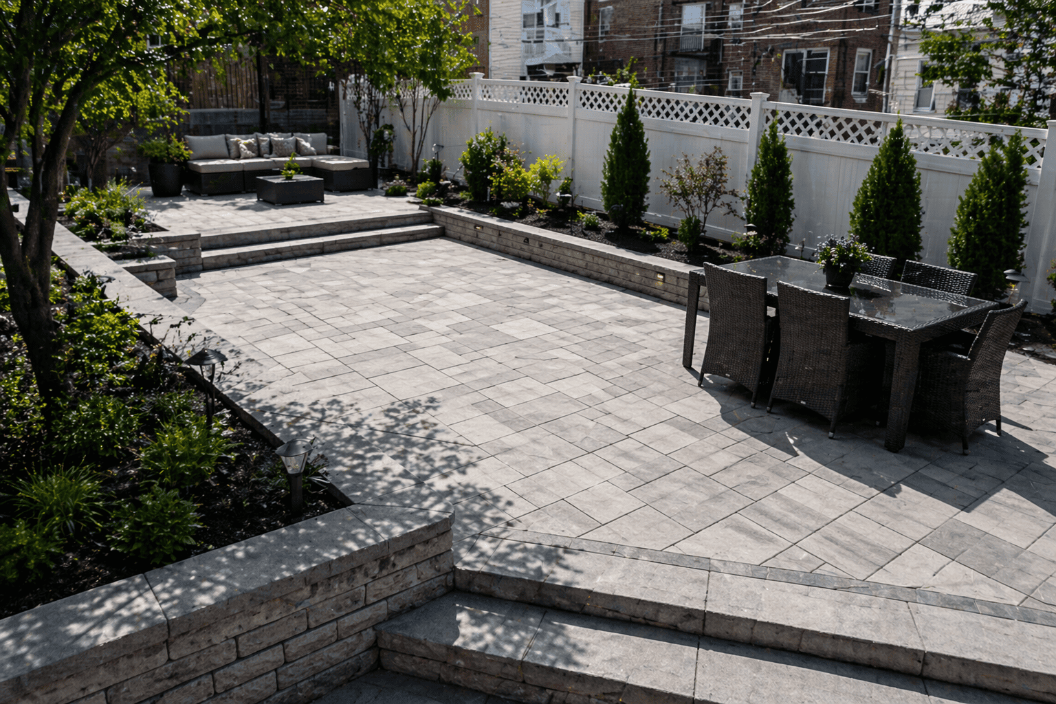 Modern backyard patio installation in Queens with large stone pavers, tiered retaining walls, built-in planting beds, and separate dining and lounge areas