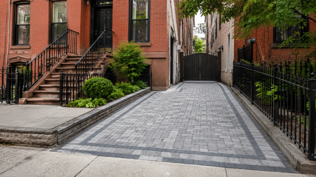 Driveway pavers in Brooklyn installed beside a brick townhouse, showing a clean gray paver driveway with a dark border, iron fencing, and landscaped curb appeal.